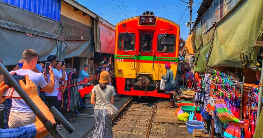 A train passing through Maeklong Railway Market as vendors and tourists stand close to the tracks.